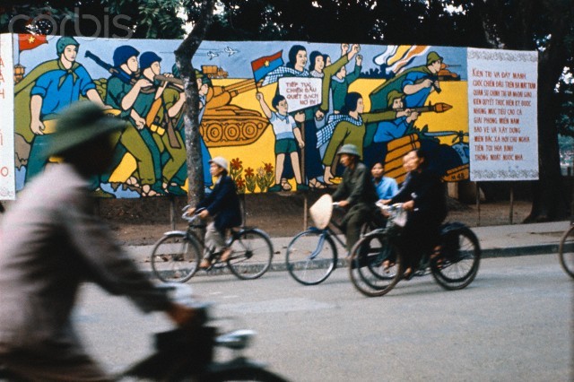 Xe đạp trên đường phố Hà Nội,bên cạnh là bức tranh cổ động lớn phản đối quân đội miền Nam Việt Nam.